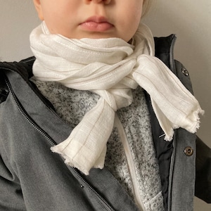 May include: A white linen scarf with thin gold stripes is tied around the neck of a child wearing a grey jacket.