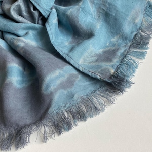 Puede incluir: Bufanda azul y gris teñida con un borde con flecos. La bufanda está hecha de una tela suave y tiene un patrón similar a una acuarela. La bufanda está doblada y drapeada, mostrando la textura y las variaciones de color.