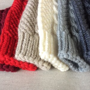 K&ouml;nnte beinhalten: Ein Stapel von sechs gestrickten M&uuml;tzen in verschiedenen Farben, darunter Burgunderrot, Rot, Beige, Wei&szlig;, Grau und Dunkelgrau. Die M&uuml;tzen sind alle mit einem Zopfmuster gestrickt.