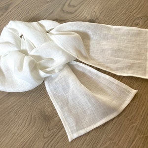 May include: A white linen scarf, elegantly draped. The scarf is made of a lightweight, textured fabric. The ends are finished with a clean, stitched hem. The scarf is a neutral accessory.