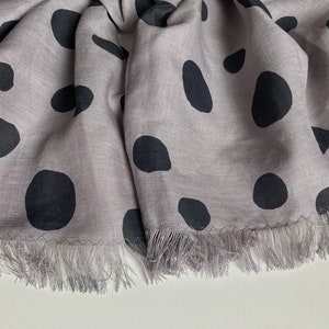 Puede incluir: Una bufanda lila con grandes lunares negros de forma irregular. La bufanda tiene un borde con flecos y está hecha de una tela suave y fluida. Un accesorio elegante.