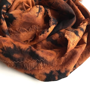 Może przedstawiać: Zbliżenie na szalik w kolorze rdzy z czarnymi wzorami tie-dye. Szalik ma postrzępioną krawędź i szyty brzeg. Tkanina jest miękka i wydaje się lekka.