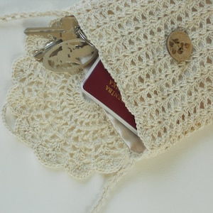 May include: A cream-coloured crocheted bag with a scalloped edge, containing keys and a passport. The bag features a button closure and a long strap. The open weave of the crochet reveals the contents. A stylish and practical accessory.