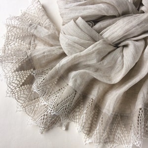 Può includere: Una sciarpa di lino beige con un delicato bordo in pizzo bianco. La sciarpa è morbida e fluida, e il pizzo aggiunge un tocco di eleganza.