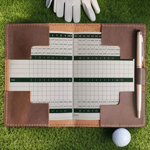 May include: A brown leather golf score card holder with a pen, scorecards, a golf ball, and a golf glove. The scorecard has numbers and spaces for recording scores. The pen is silver.