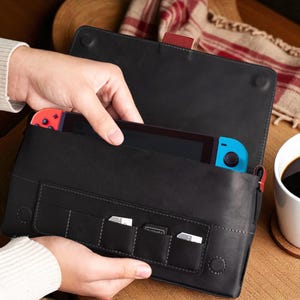 Może przedstawiać: Czarny skórzany futerał do przenoszenia konsoli do gier, z widocznymi czerwonymi i niebieskimi kontrolerami. Etui posiada zapięcie na klapę i kilka małych kieszeni na kartridże do gier. Ręka wkłada konsolę do środka.