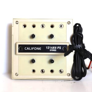 May include: A vintage Califone 1214AV-PS stereo audio selector in a cream-colored case. It features multiple input jacks, black knobs, and the Califone logo. A black cable is coiled to the side with a red and white tag.