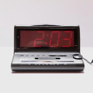 May include: A black digital alarm clock displaying the time 2:03 in red numerals. The clock has several buttons and a radio dial. A black power cord extends from the side.