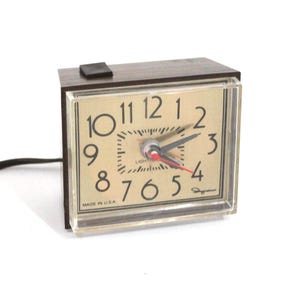 May include: A rectangular electric alarm clock with a dark brown wood-grain finish case and a clear plastic front cover. The clock face is gold-toned with black Arabic numerals, black hour and minute hands, and a red second hand. Text reads MADE IN U.S.A.
