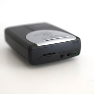 Puede incluir: Un reproductor de casetes negro y plateado con una toma de auriculares y una perilla de control de volumen.