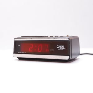 May include: A black and silver Cosmo Time alarm clock displaying the time 2:07 in red digital numbers. The clock has a rectangular shape with a silver trim and a black power cord.
