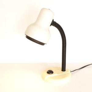 May include: A desk lamp featuring a cream-colored dome-shaped shade and a matching circular base. The lamp has a flexible black neck and a small black power switch located on the top of the base. A thin black power cord extends from the back.