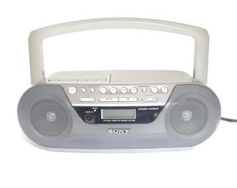 May include: A silver Sony CD radio cassette recorder with a top-mounted handle, dual front-facing speakers, a central digital display, and various control buttons. The unit features a CD-R/RW playback label and an audio input jack on the front panel.