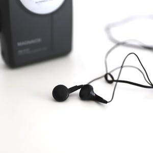 Puede incluir: Un reproductor de casetes negro con una etiqueta blanca y auriculares negros con un cable negro.