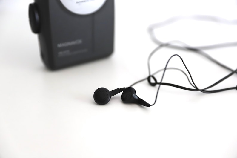 Puede incluir: Un reproductor de casetes negro con una etiqueta blanca y auriculares negros con un cable negro.