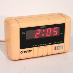 May include: A rectangular peach-colored Conair digital alarm clock with a black display panel showing the time 2:05 in bright red LED numbers. The device features a snooze button, time and alarm set controls, and an on-off alarm switch.