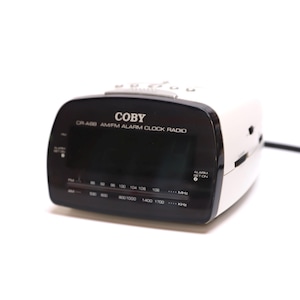 May include: A white and black Coby CR-488 AM/FM alarm clock radio. The clock has a black display with radio frequency indicators and the word "COBY" printed on the top. A black power cord extends from the side.