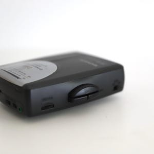 Puede incluir: Un reproductor de casetes negro con una cara plateada y botones. El reproductor tiene una toma de auriculares y una perilla de control de volumen.