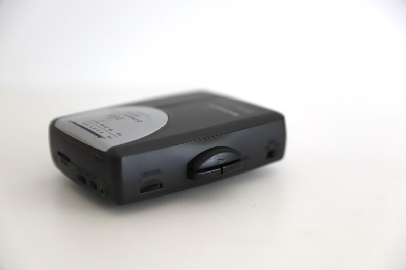 Puede incluir: Un reproductor de casetes negro con una cara plateada y botones. El reproductor tiene una toma de auriculares y una perilla de control de volumen.
