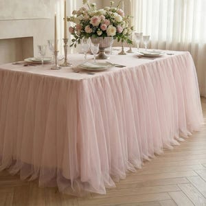 May include: A table set for a celebration, draped with a light pink tulle tablecloth. The table is set with plates, glasses, and cutlery. A large floral arrangement of white and pink flowers sits in a silver vase, with candles.