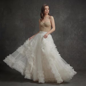 Puede incluir: Un top corsé beige sin tirantes combinado con una voluminosa falda de tul blanco. La falda presenta múltiples capas de tela con volantes, creando una silueta fluida y elegante. Adecuado para ocasiones especiales.