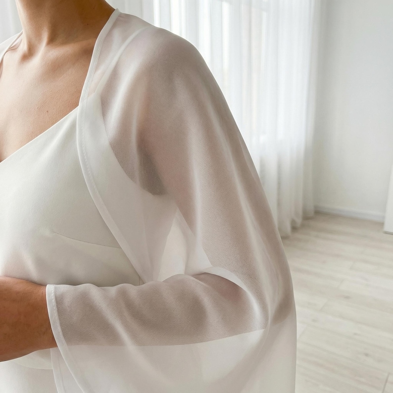 May include: A sheer white shawl draped over a white dress. The shawl has long sleeves and a delicate, flowing appearance. The fabric is lightweight and translucent, creating a soft, elegant look. The style is suitable for formal events.