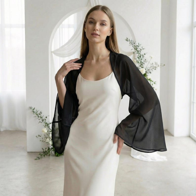 May include: A woman wearing a black sheer bolero over a white slip dress. The bolero has long, flowing sleeves. The background is a bright, white room with floral arrangements and a large arched doorway.