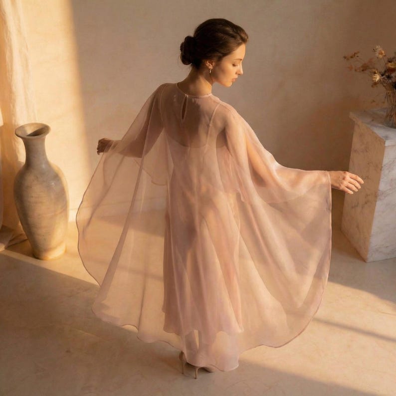 May include: A flowing, blush-colored dress with a sheer cape overlay. The dress has a simple silhouette and a high neckline. The cape drapes elegantly, creating a soft, ethereal look. The fabric appears lightweight and airy, perfect for a special occasion.