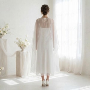 May include: A white wedding dress with a sheer cape. The dress has a simple silhouette and is paired with white heels. The cape drapes over the shoulders and down the back, creating an elegant look. The background is a bright, white room.