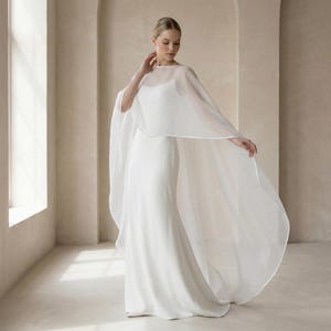 May include: A white wedding dress with a sheer cape. The dress is a simple, elegant design with a fitted silhouette. The cape is made of a lightweight, flowing fabric that drapes over the shoulders and arms. The overall look is classic and romantic.