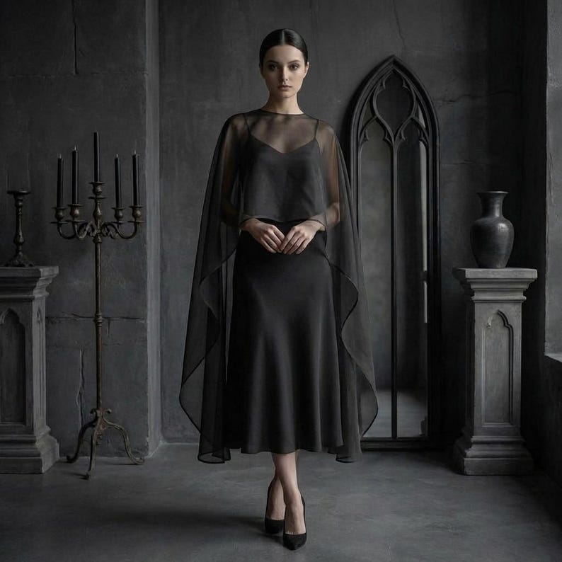 May include: A woman in a black dress and sheer cape stands in a dark room. The dress is a midi-length, and the cape drapes over the shoulders. The room features a gothic mirror, a candelabra, and a decorative vase.