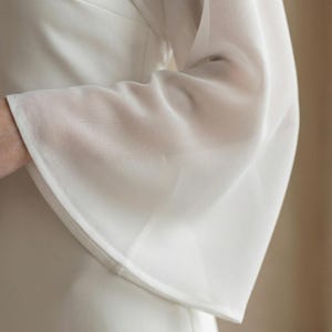 May include: Close-up of a white, long-sleeved garment with a flared sleeve. The fabric appears to be lightweight and slightly sheer, with a subtle texture. The sleeve has a clean, finished hem.