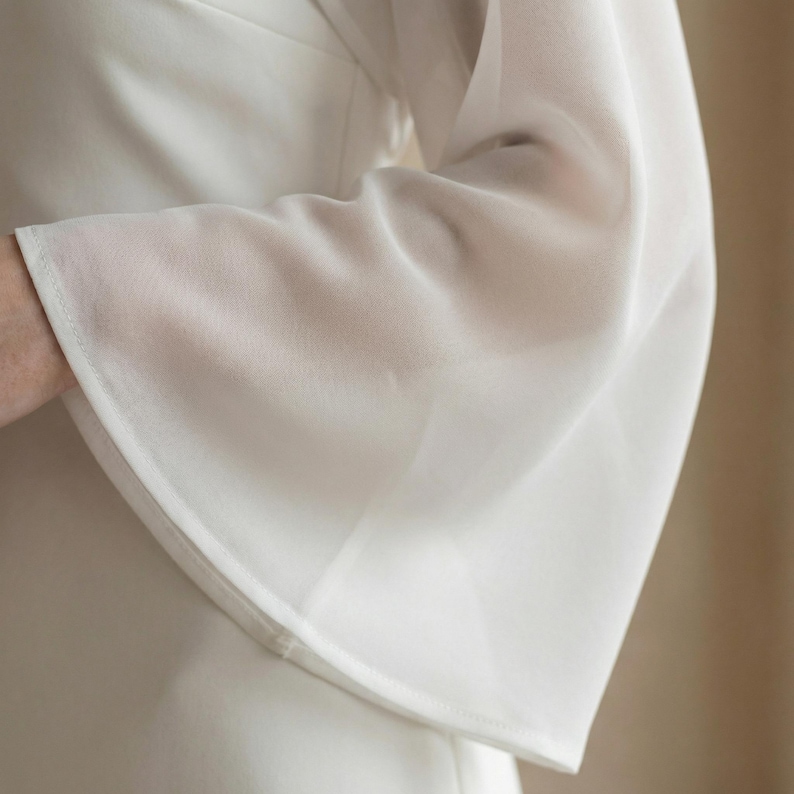 May include: Close-up of a white, long-sleeved garment with a flared sleeve. The fabric appears to be lightweight and slightly sheer, with a subtle texture. The sleeve has a clean, finished hem.
