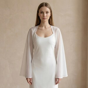 May include: A white, floor-length dress with a scoop neckline is paired with a sheer, long-sleeved bolero. The bolero has a square neckline and wide sleeves. The outfit is simple and elegant, perfect for a special occasion.