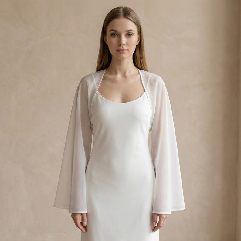 May include: A white, floor-length dress with a scoop neckline is paired with a sheer, long-sleeved bolero. The bolero has a square neckline and wide sleeves. The outfit is simple and elegant, perfect for a special occasion.