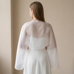 May include: A white, sheer, long-sleeved bridal capelet over a white dress. The capelet has a delicate, flowing design with wide sleeves. The dress has a simple, elegant silhouette. The background is a neutral beige.