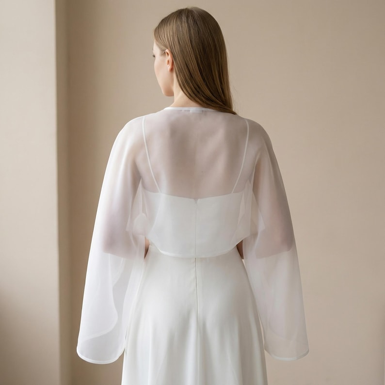 May include: A white, sheer, long-sleeved bridal capelet over a white dress. The capelet has a delicate, flowing design with wide sleeves. The dress has a simple, elegant silhouette. The background is a neutral beige.