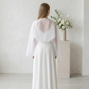 May include: A white bridal ensemble featuring a long, flowing skirt and a sheer, long-sleeved cape. The cape drapes over the shoulders, creating an elegant silhouette. The outfit is set against a neutral background, with a vase of white flowers.
