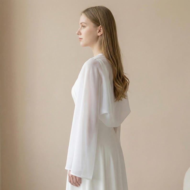 May include: A white, flowing dress with long sleeves and a sheer cape overlay. The dress has a simple, elegant design, perfect for a wedding or special occasion. The fabric appears lightweight and airy, creating a graceful silhouette.