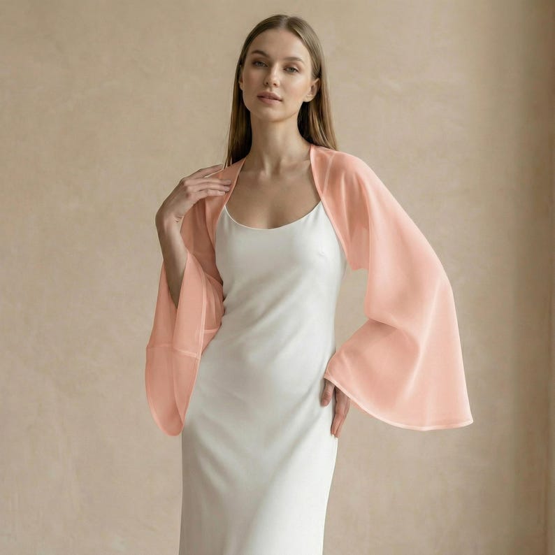May include: A peach-colored, sheer bolero with wide, flowing sleeves. The bolero is worn over a white, sleeveless dress. The fabric appears lightweight and airy, creating a soft, elegant look. The outfit is suitable for special occasions.