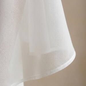 May include: Close-up of a white, semi-sheer fabric with a slightly textured surface. The fabric is draped, creating soft folds and shadows. The edge is finished with a delicate, stitched hem.