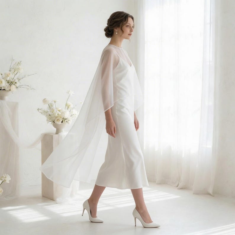 May include: A white wedding dress with a sheer cape overlay. The dress is a simple, elegant design with a midi length. The model is wearing white high heels. The background is a bright white room with a large window.