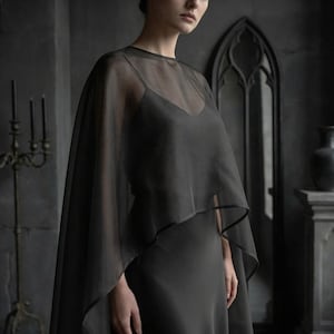 May include: A dark gray, sheer cape with a black trim, worn over a matching slip dress. The cape drapes over the shoulders, creating a flowing silhouette. The background features a dark interior with gothic architectural elements.