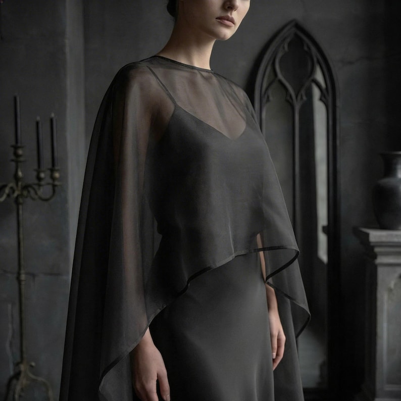 May include: A dark gray, sheer cape with a black trim, worn over a matching slip dress. The cape drapes over the shoulders, creating a flowing silhouette. The background features a dark interior with gothic architectural elements.