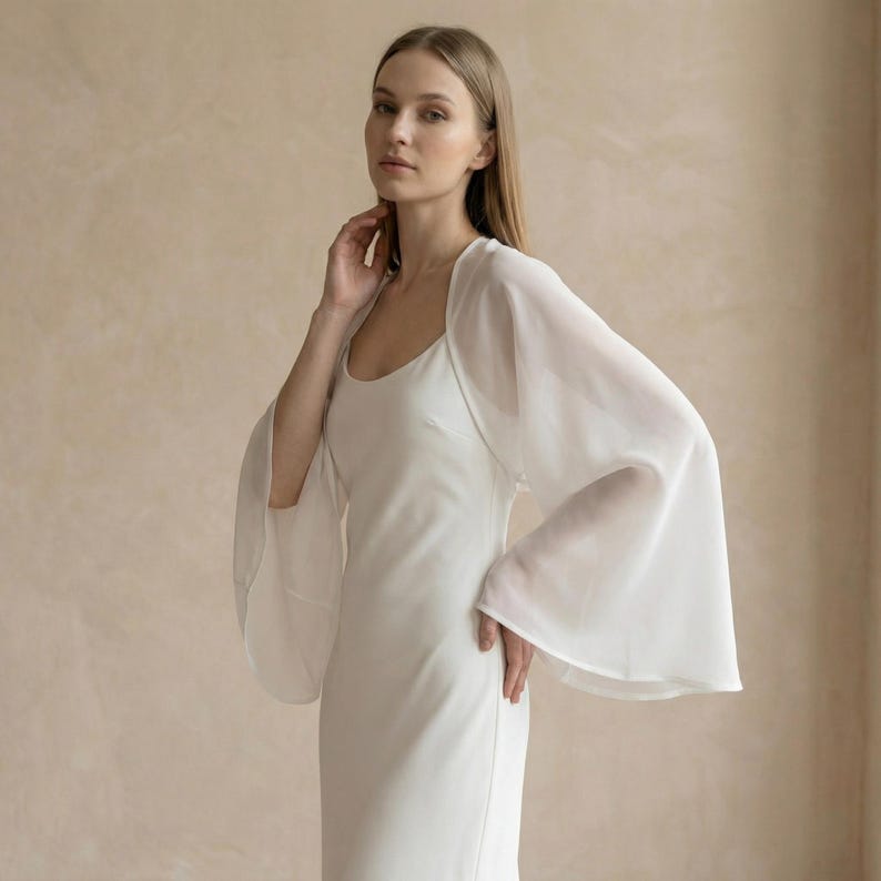 May include: A white, floor-length dress with a scoop neckline. The dress is paired with a sheer, white, bell-sleeved bolero. The model is standing in front of a neutral-toned background.