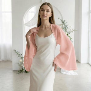 May include: A woman wearing a light peach-colored chiffon bolero with wide sleeves over a white dress. The bolero is sheer and has a soft, flowing appearance. The background is a light-filled room with floral accents.