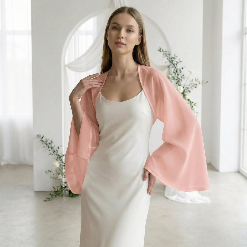 May include: A woman wearing a light peach-colored chiffon bolero with wide sleeves over a white dress. The bolero is sheer and has a soft, flowing appearance. The background is a light-filled room with floral accents.