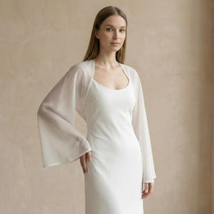 May include: A white, sleeveless dress with a scoop neckline. A sheer, long-sleeved bolero in a matching white color is worn over the dress. The bolero has a square neckline and wide, flowing sleeves. The background is a solid, neutral color.
