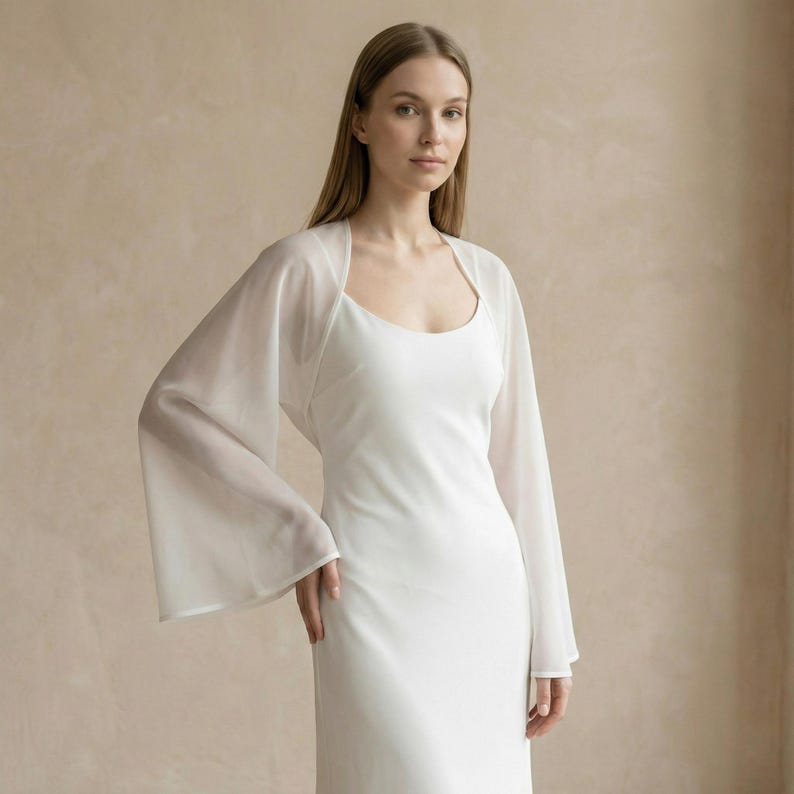 May include: A white, sleeveless dress with a scoop neckline. A sheer, long-sleeved bolero in a matching white color is worn over the dress. The bolero has a square neckline and wide, flowing sleeves. The background is a solid, neutral color.