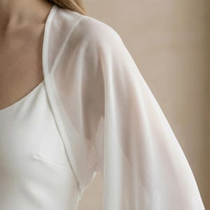 May include: A white, sheer, long-sleeved bolero. The bolero is made of a lightweight, flowing fabric. The garment is worn over a white dress. The bolero has a delicate, elegant appearance, suitable for formal occasions.